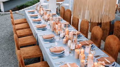 Long dining table set with candles, glasses, plates, and brown napkins, surrounded by brown chairs.