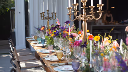 Long wooden table set outdoors with colorful flowers, glassware, plates, and gold candleholders.