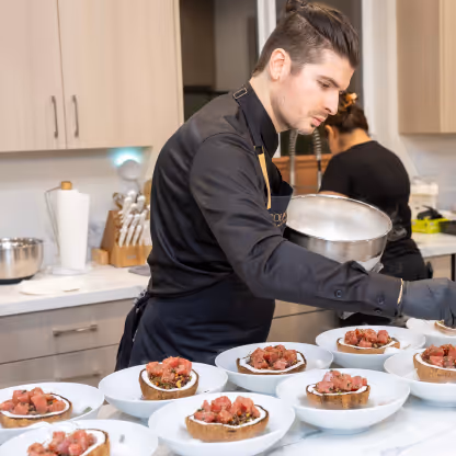 Chef in black outfit plating multiple bowls of food in a modern kitchen setting.