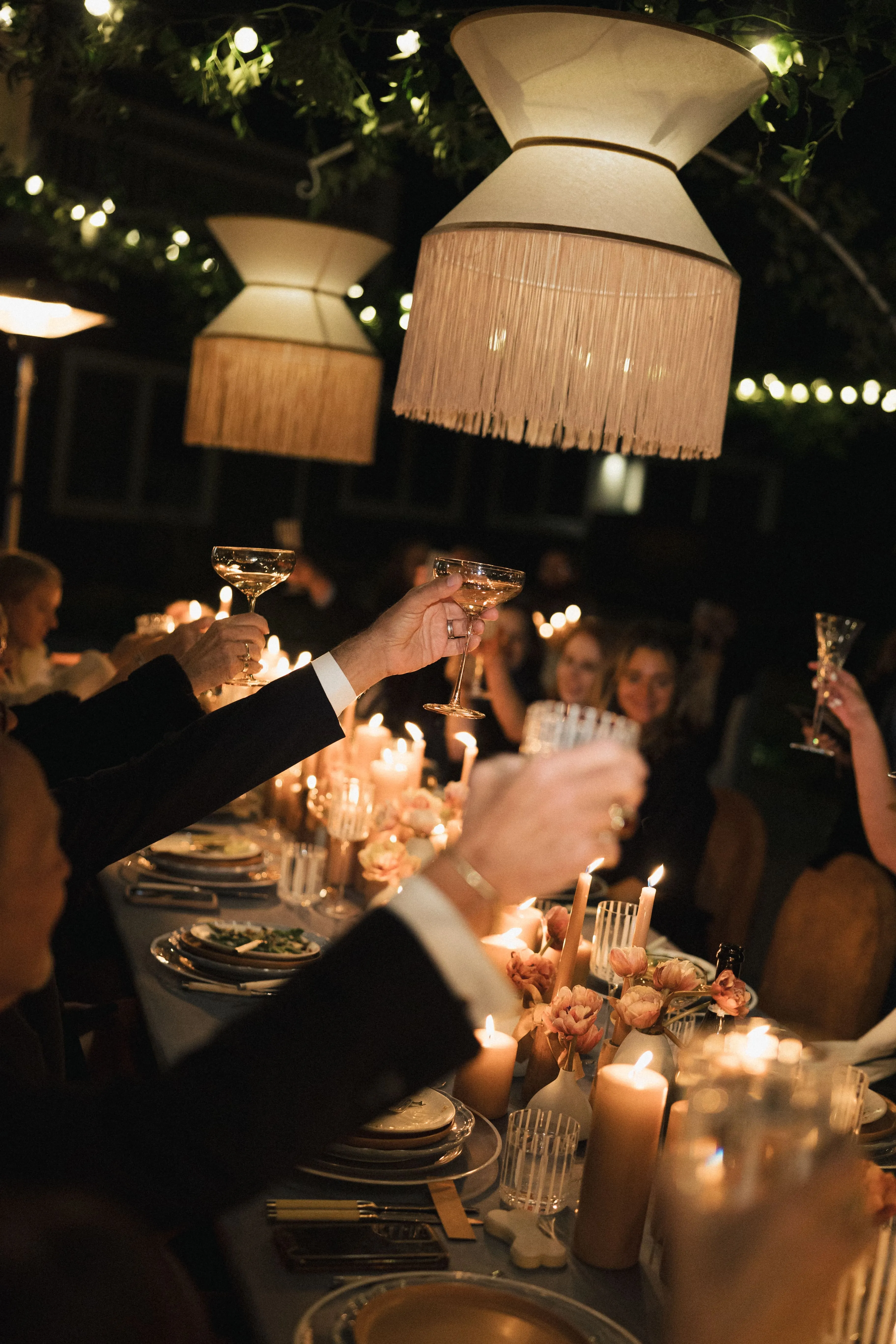 People raising glasses in a candlelit dinner setting under hanging lamps and string lights.