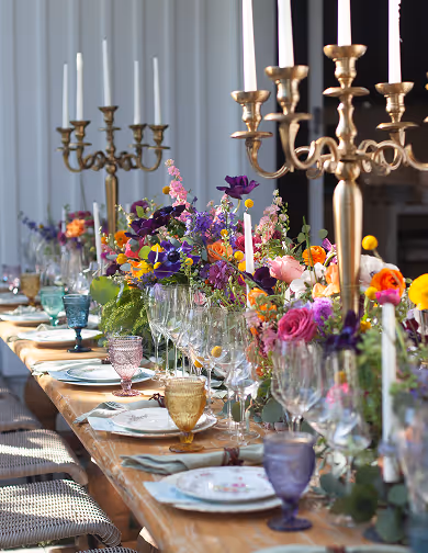Long wooden table set with colorful flowers, glassware, plates, and gold candle holders with lit candles.