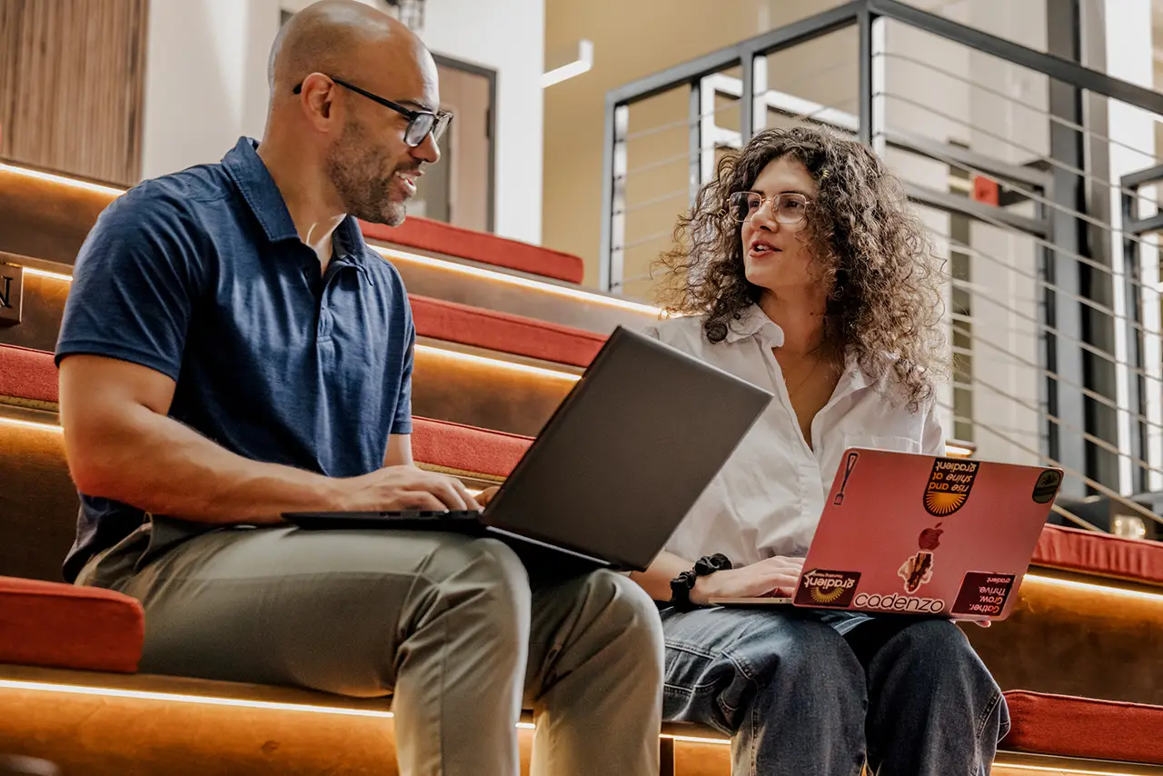 Two people sitting on carpeted steps working on laptops and talking in a modern office space.