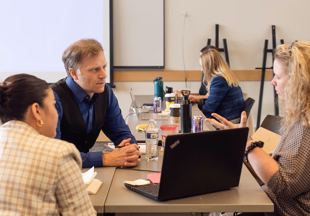 A group of colleagues seated around a table with laptops during a working meeting in a conference room.