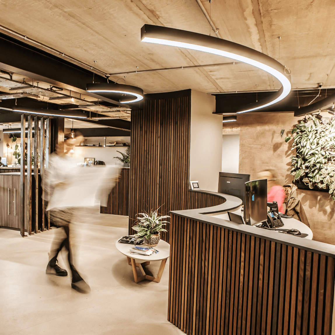 Modern office reception with curved wood desk, circular lights, plants, and blurred people moving inside.