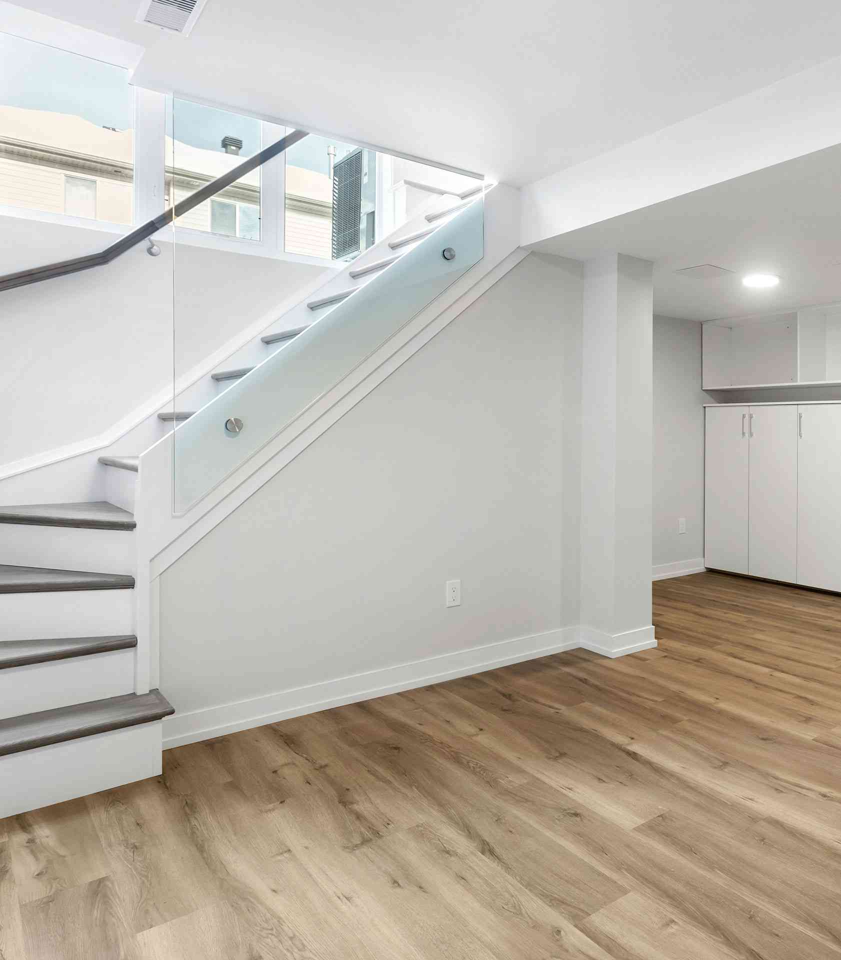 Finished basement with glass-railed stairs, wood-look flooring and storage cabinets, highlighting waterproofed space