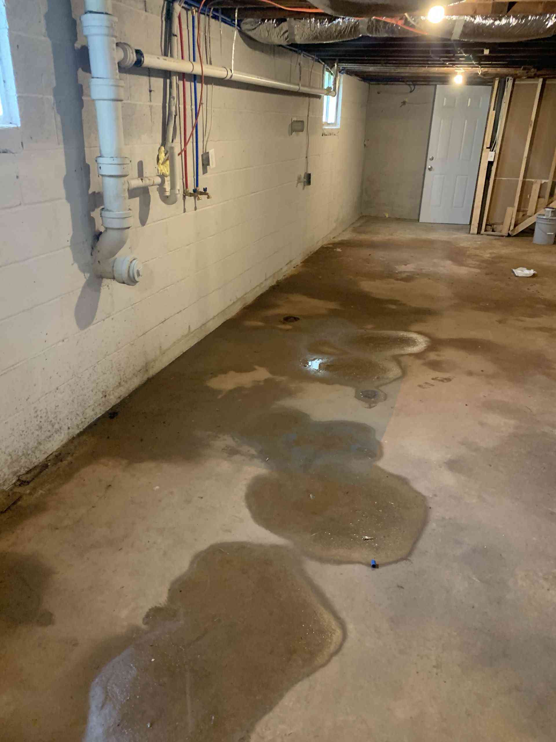 Basement concrete floor with multiple water puddles, exposed drain and wall plumbing showing moisture intrusion