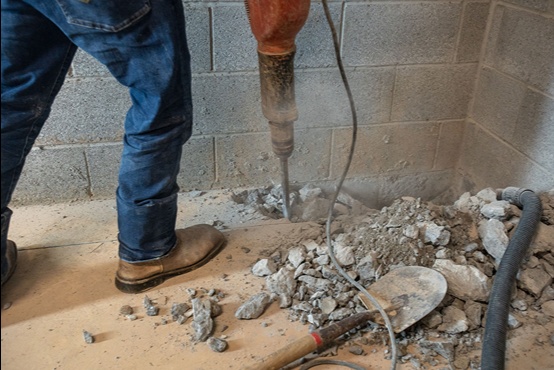 Technician using jackhammer to break concrete floor next to cinderblock foundation for interior drain/sump work