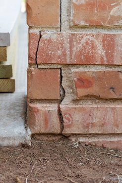 Vertical crack through brick foundation corner and mortar beside slab, showing foundation settlement damage