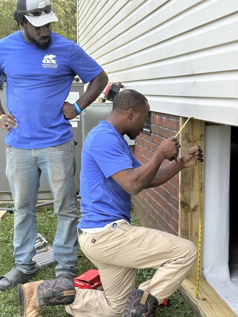 Two Ox technicians measuring a crawl-space access between brick and siding with a tape measure for foundation repair
