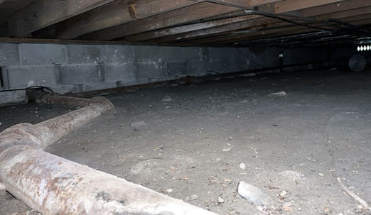 Dirt crawl space with rusted pipe, exposed joists and cinderblock wall showing need for encapsulation or repair.