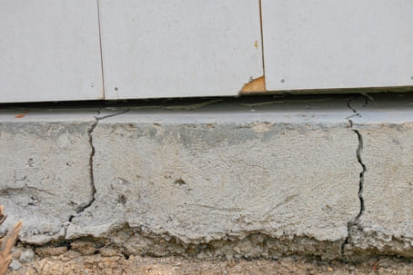 Concrete foundation with vertical cracks and a gap under siding, showing settlement and need for foundation repair