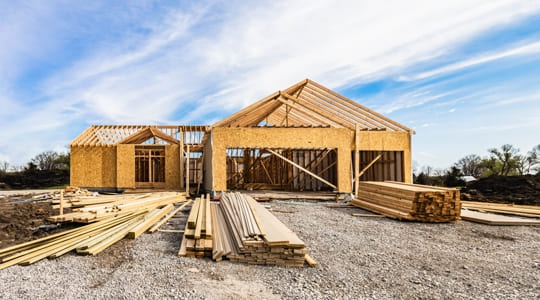 Wood-framed house under construction with stacked lumber and exposed foundation highlighting structural framing