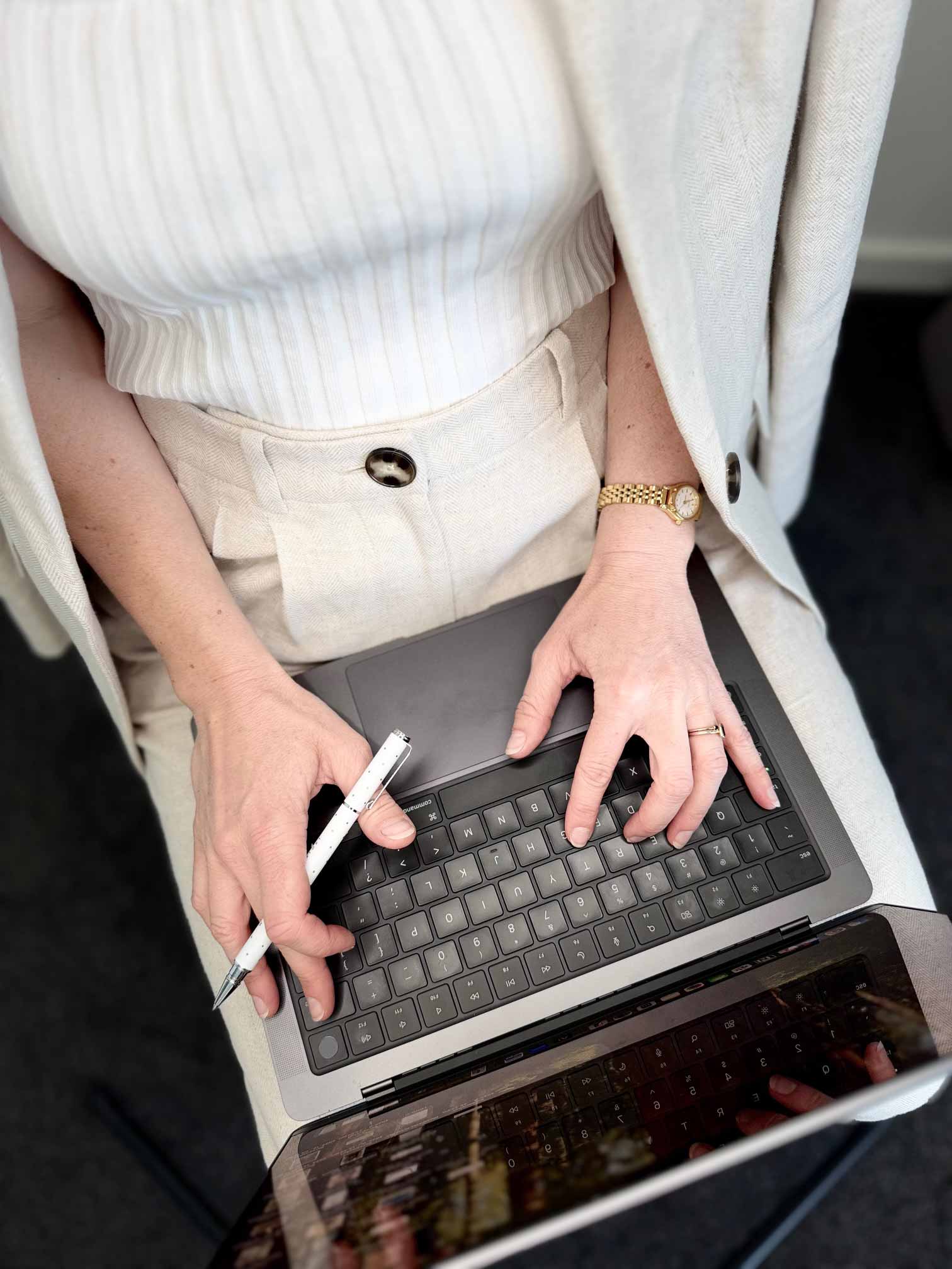 POV point of view of a woman typing on a macbook laptop with a white pen in hand, wearing a knitted top and light beige blazer and pants
