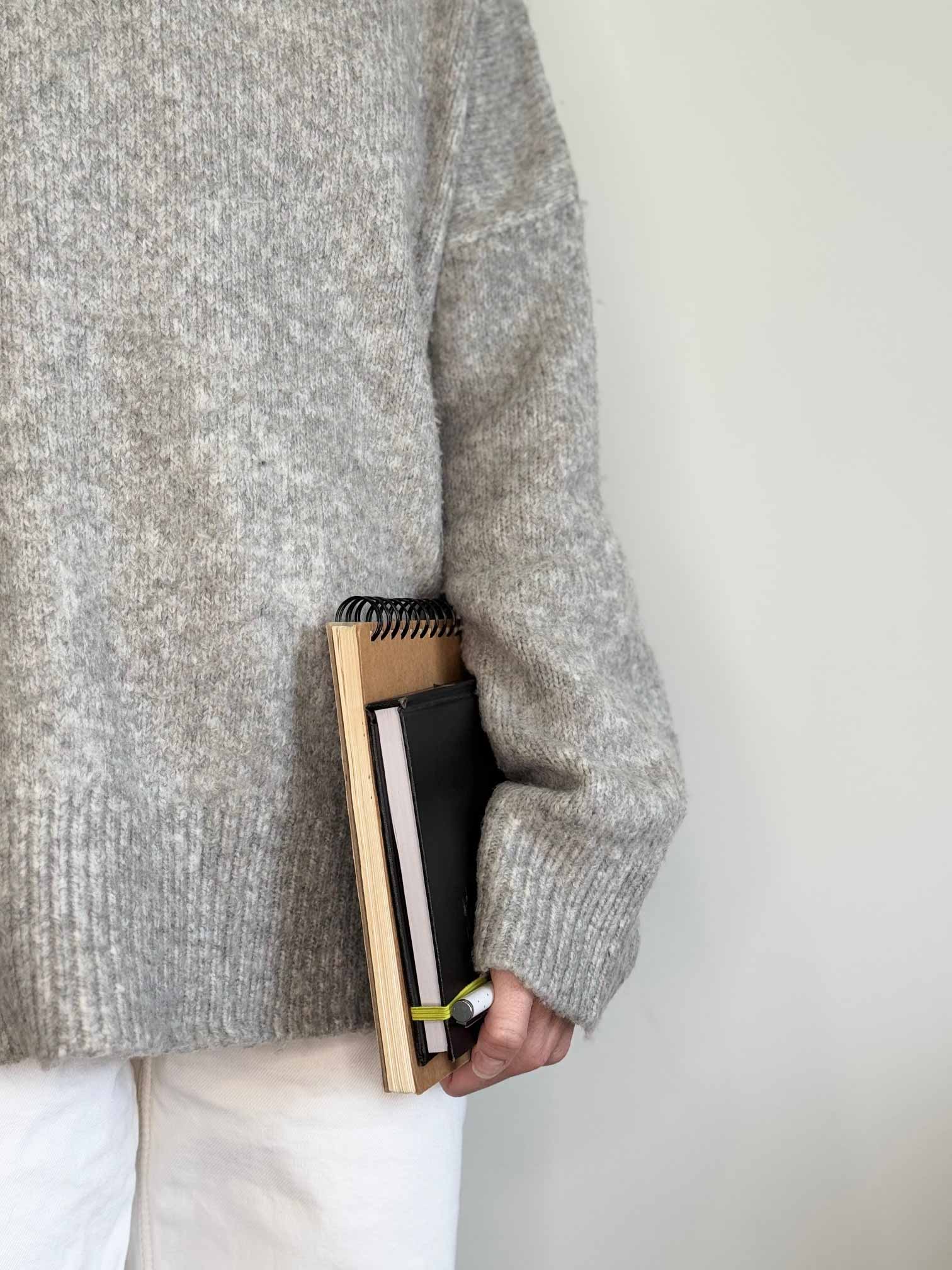 A person in a grey knitted jumper and white jeans holding a tan sketchpad, black diary and white pen