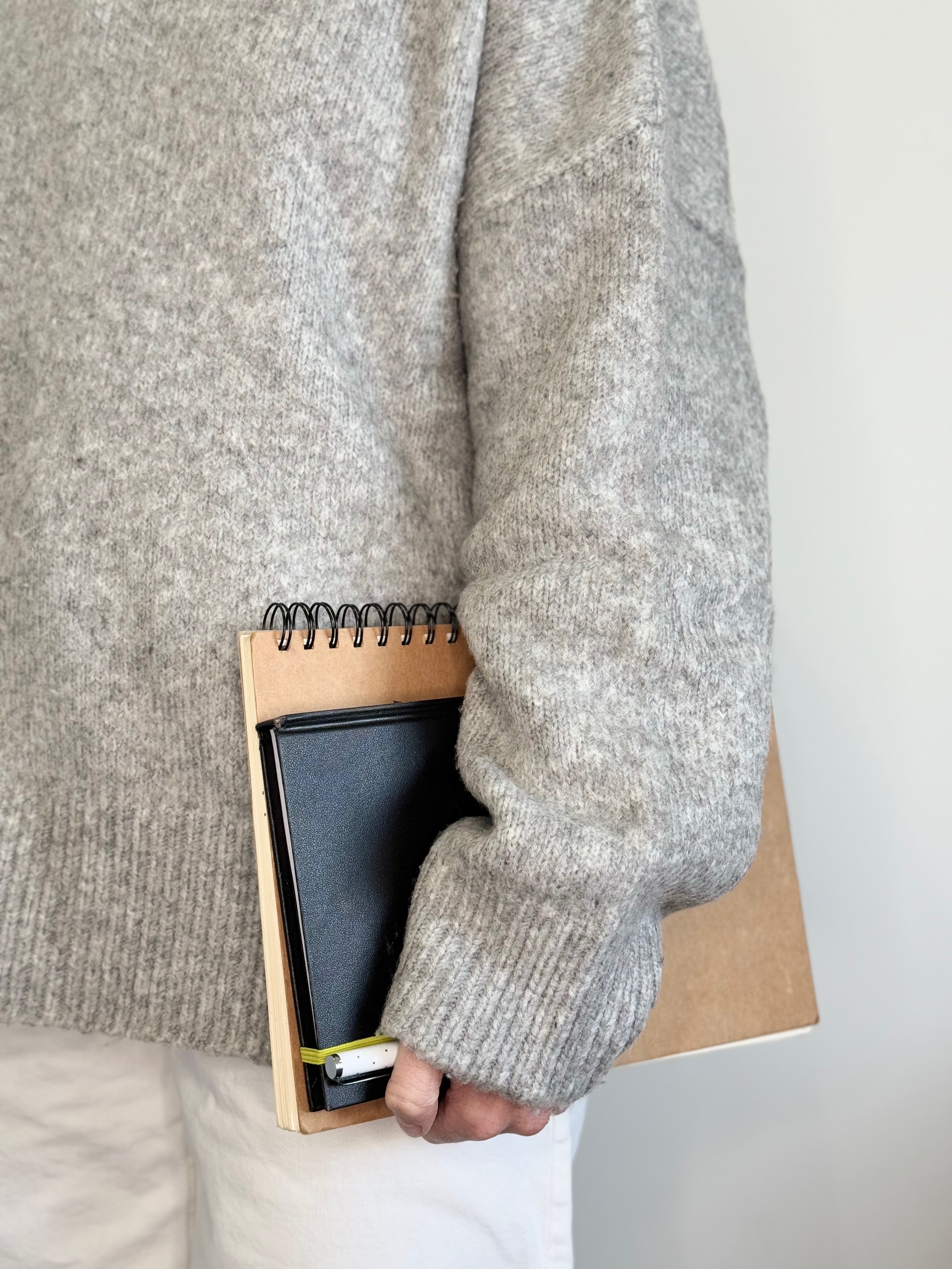 Cropped image of a person in a grey knitted jumper and white jeans carrying a tan sketch book, black diary and white pen
