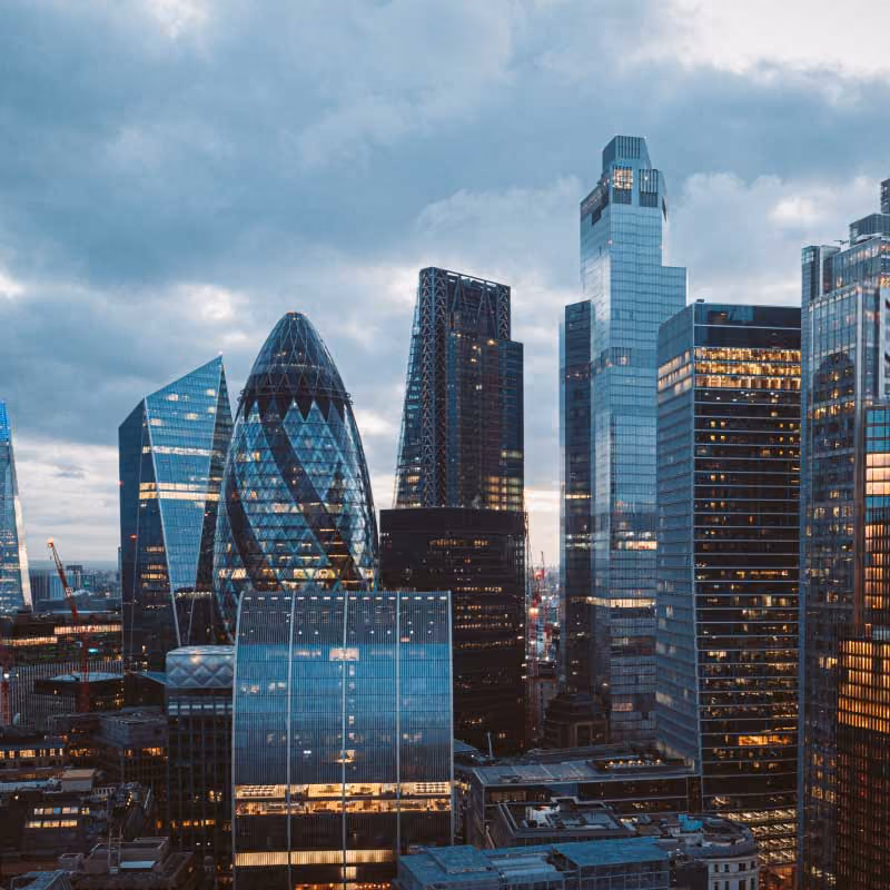 A cityscape at dusk with illuminated modern skyscrapers against a cloudy sky in the Uk.