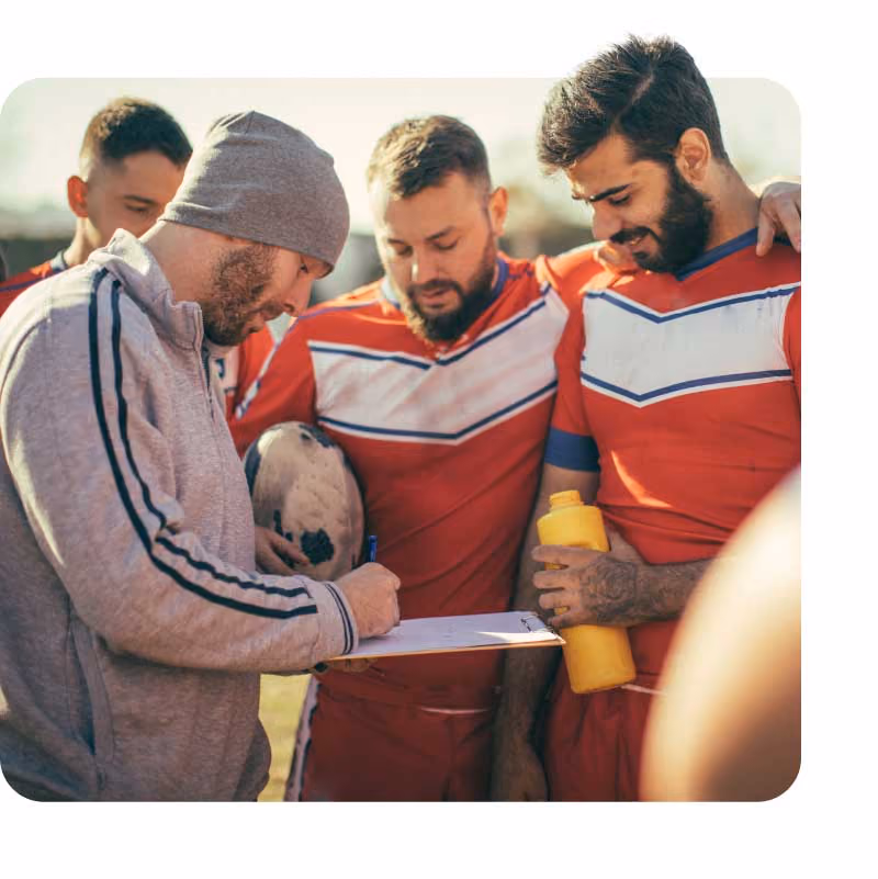 Group of rugby players in red jerseys listening to their coach holding a clipboard.