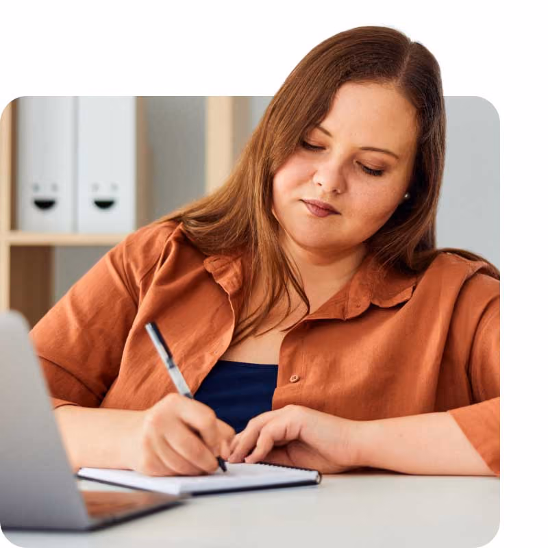 Person in an orange shirt writing in a notebook at a desk with a laptop.