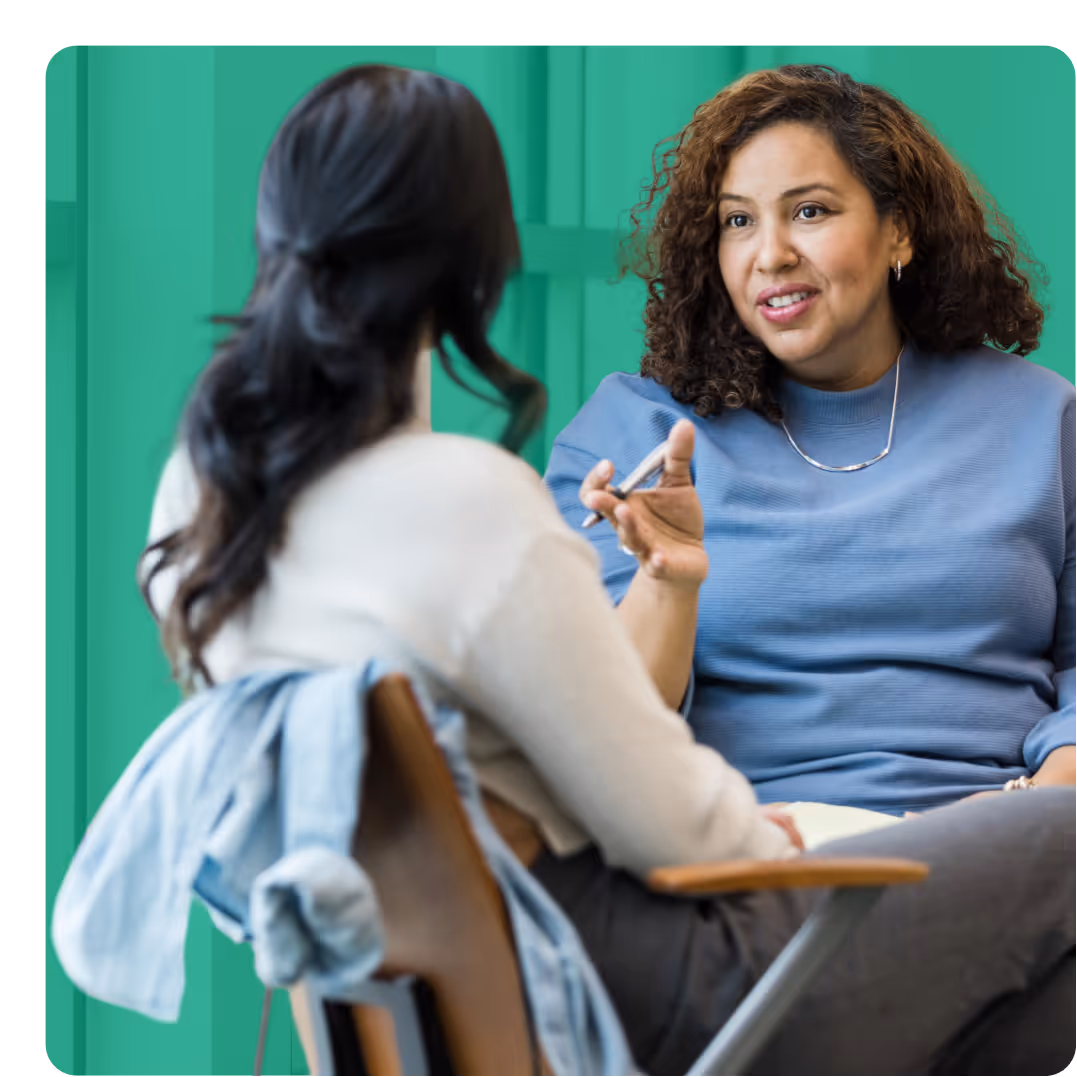 Two women engaged in a conversation, one with curly hair wearing a blue top and the other with long dark hair in a white top sitting on chairs.