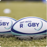 Two rugby balls on grass with text 'GET INTO RUGBY' in blue letters.