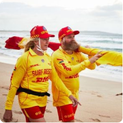 Two surf lifesavers in yellow and red uniforms carrying a rescue board on a beach.