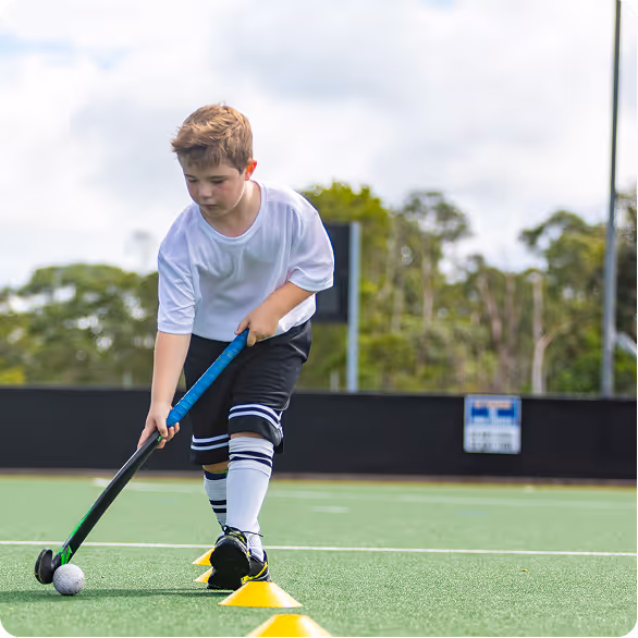 Young boy practicing field hockey with a stick and ball on a sports field marked with yellow cones.