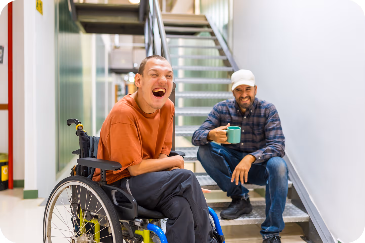 Man in wheelchair and another man sitting on stairs, both laughing and enjoying a moment.
