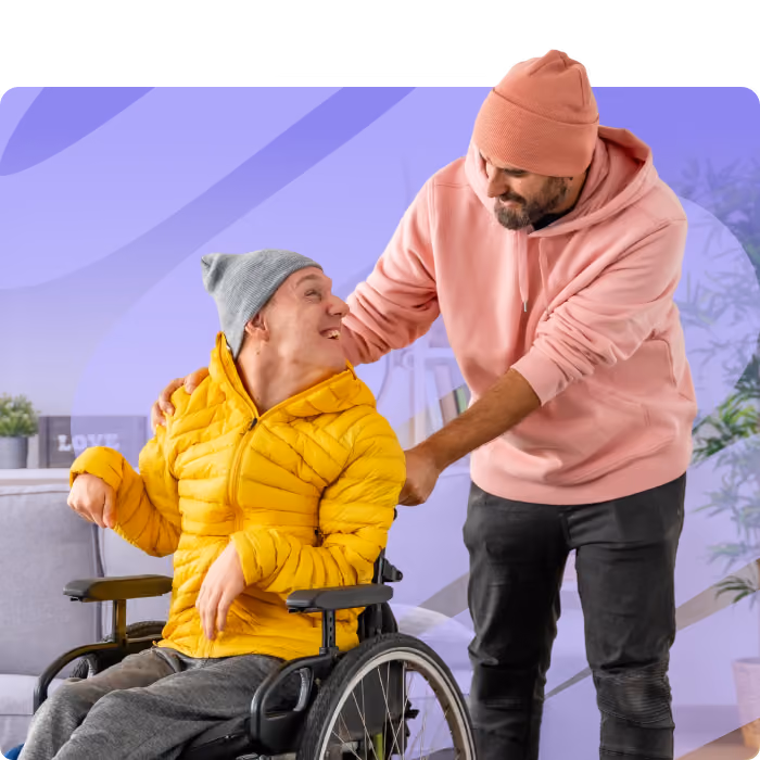Man in a pink hoodie assisting a smiling man in a wheelchair wearing a yellow jacket and gray beanie.
