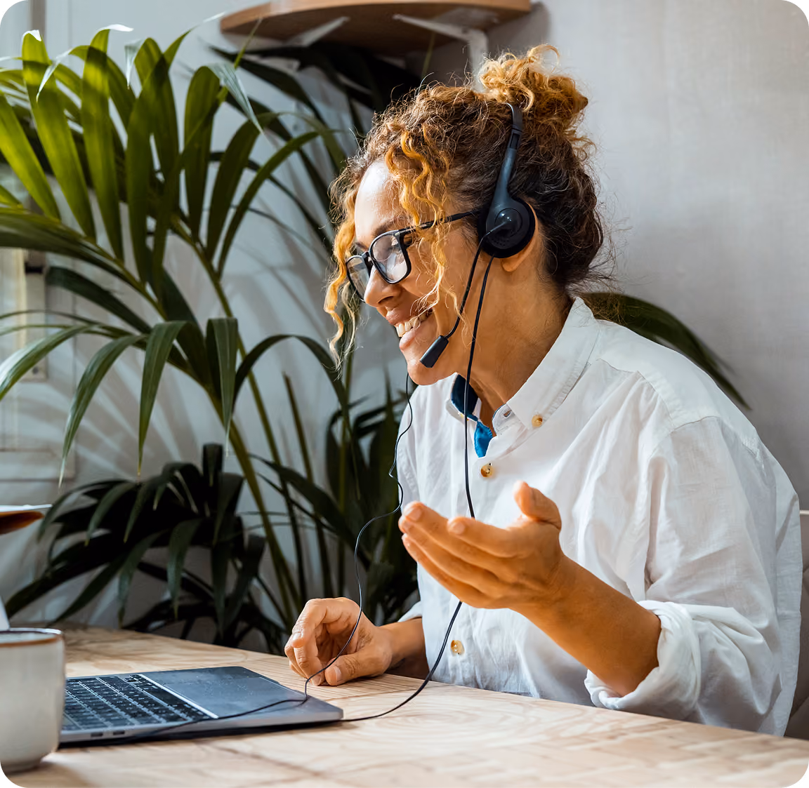 Smiling woman with curly hair and glasses wearing a headset while using a laptop at a wooden table with green plants in the background.
