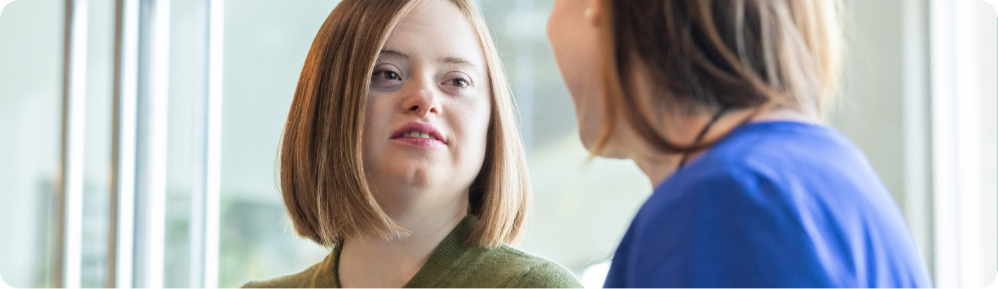 A support worker holding a tablet talks with a young woman with Down syndrome, who has her hand on her chest as she speaks.