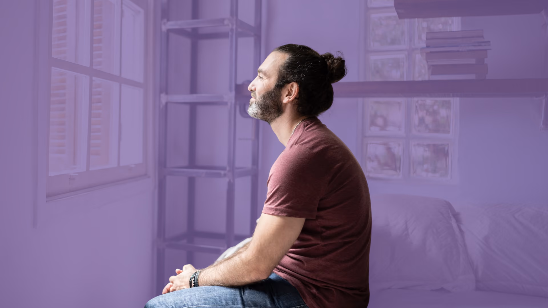 A man sits on the edge of a bed, looking out a window.