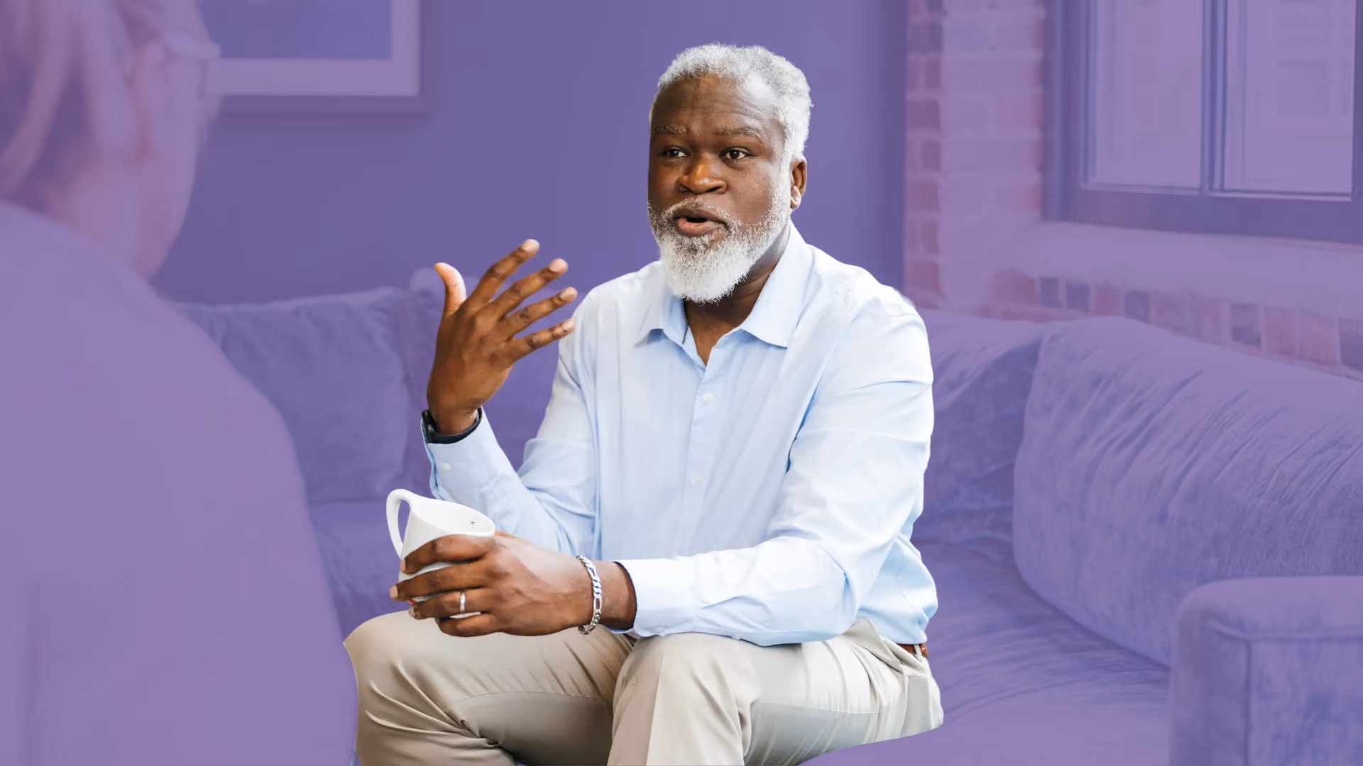 An older man holding a mug talks and gestures while sitting on a couch across from another person, with a purple colour overlay.
