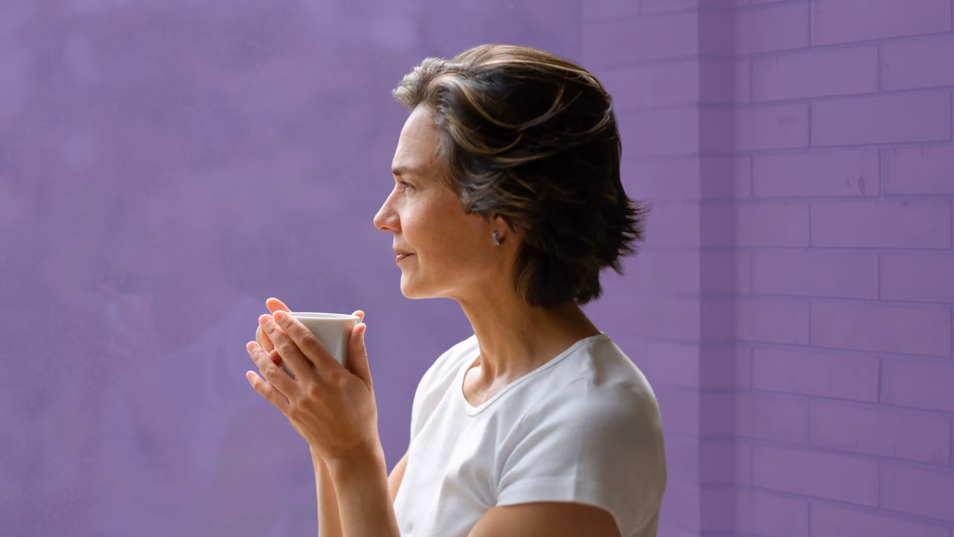 A woman holds a mug in both hands and gazes out a window, in profile, with a purple colour overlay.