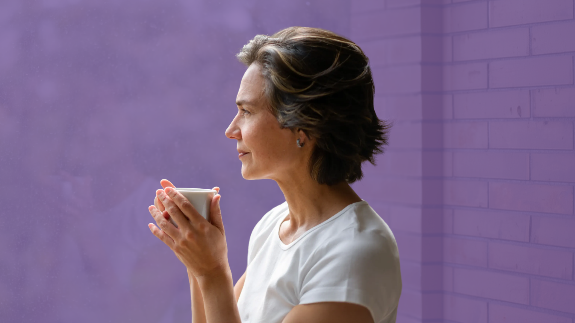 A woman holds a mug in both hands and gazes out a window, in profile, with a purple colour overlay.