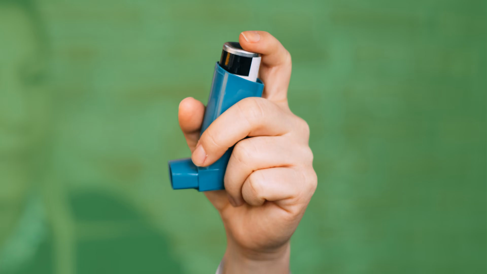 A hand holds up a blue asthma inhaler against a green background.