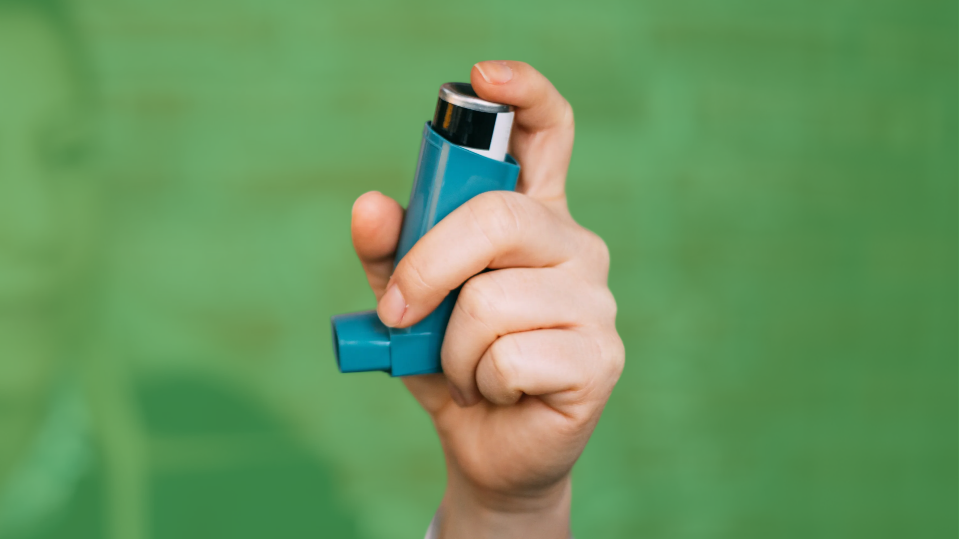 A hand holds up a blue asthma inhaler against a green background.