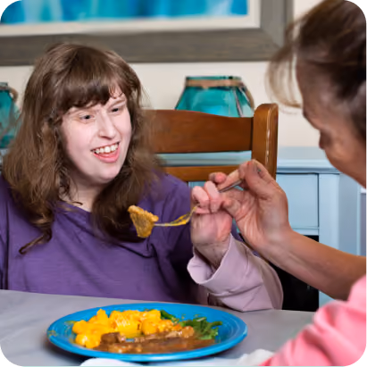 A smiling woman in a purple top is offered a spoonful of food by another person while sitting at a table.