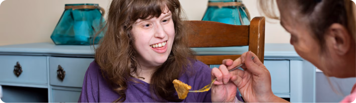 A smiling woman in a purple top is offered a spoonful of food by another person while sitting at a table.
