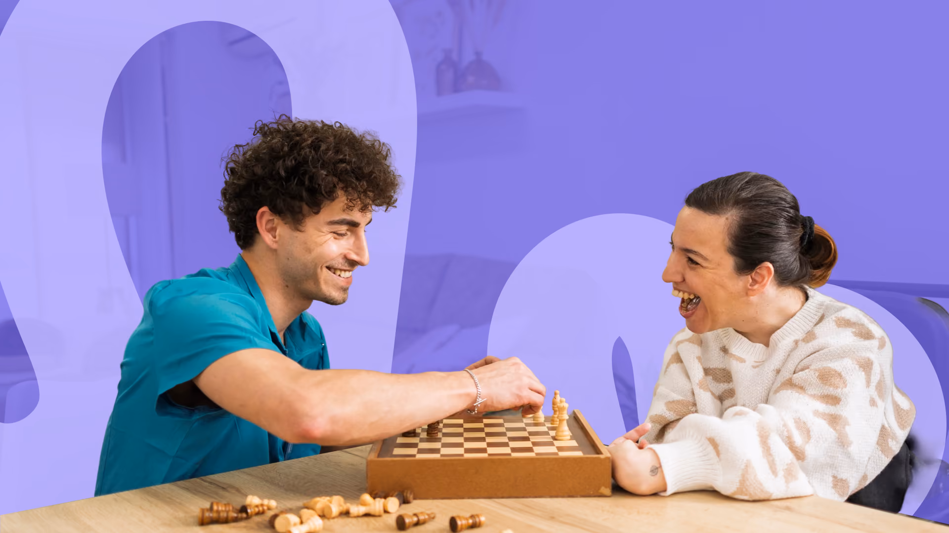 A support worker and a woman laughing together while playing a game of chess at a table.