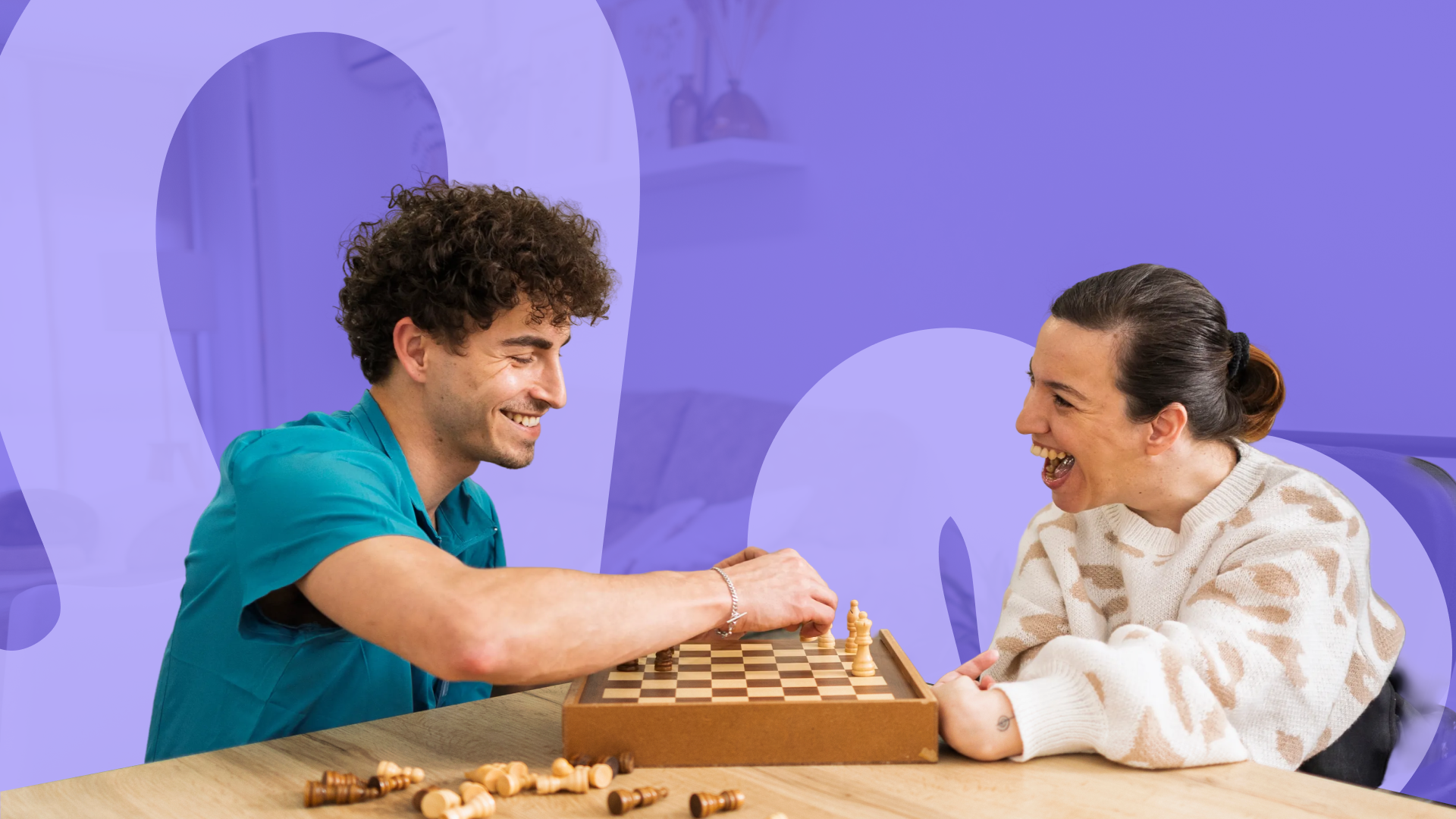 A support worker and a woman laughing together while playing a game of chess at a table.