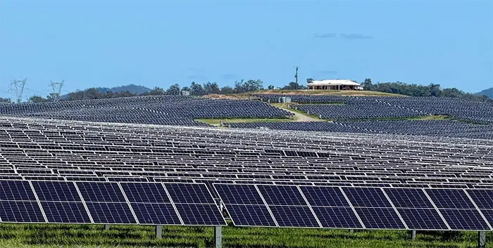 A vast field of solar panels stretching across rolling green hills under a clear blue sky, with a small building and power lines visible in the background.