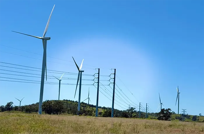 Multiple white wind turbines stand on a grassy hill alongside tall transmission towers and power lines under a bright blue sky.