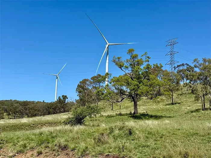 Two large wind turbines and a transmission tower rise above a grassy, tree-dotted hill under a clear blue sky.