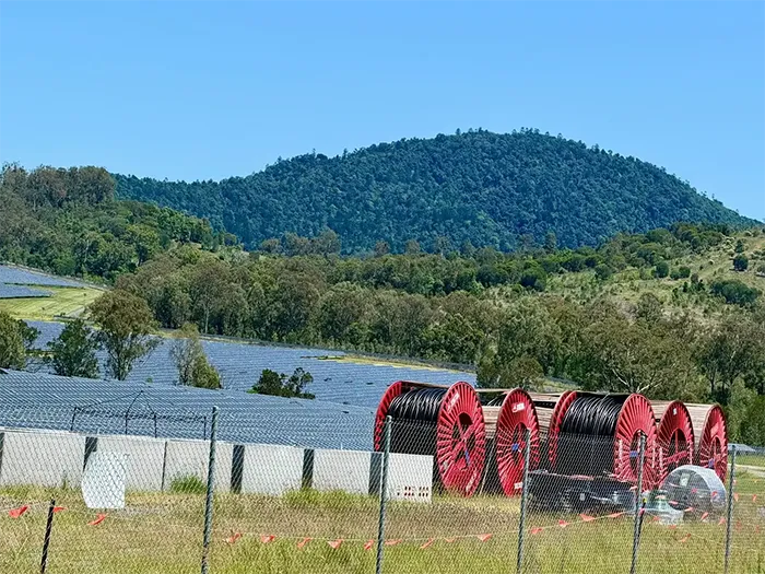 Large red industrial cable spools in the foreground of a solar farm site, with a prominent forested hill in the background.