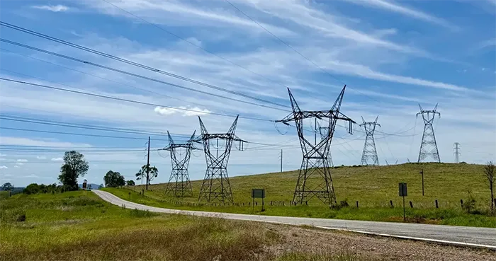 A line of tall steel lattice transmission towers carries power lines across a grassy field next to a rural road under a streaky blue sky.