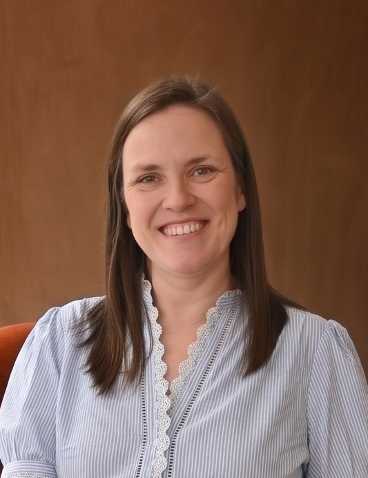 Smiling woman with straight brown hair wearing a floral top against a solid brown background.