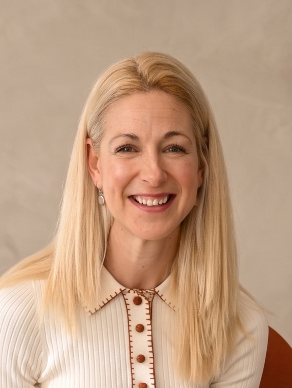 Smiling woman with long blonde hair wearing a green top standing indoors near a plant.