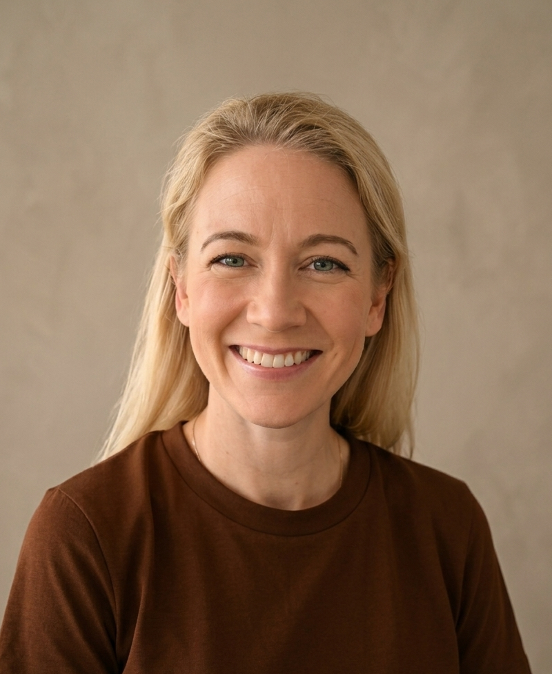 Smiling woman with blonde hair wearing a brown sweater and smartwatch sitting indoors.