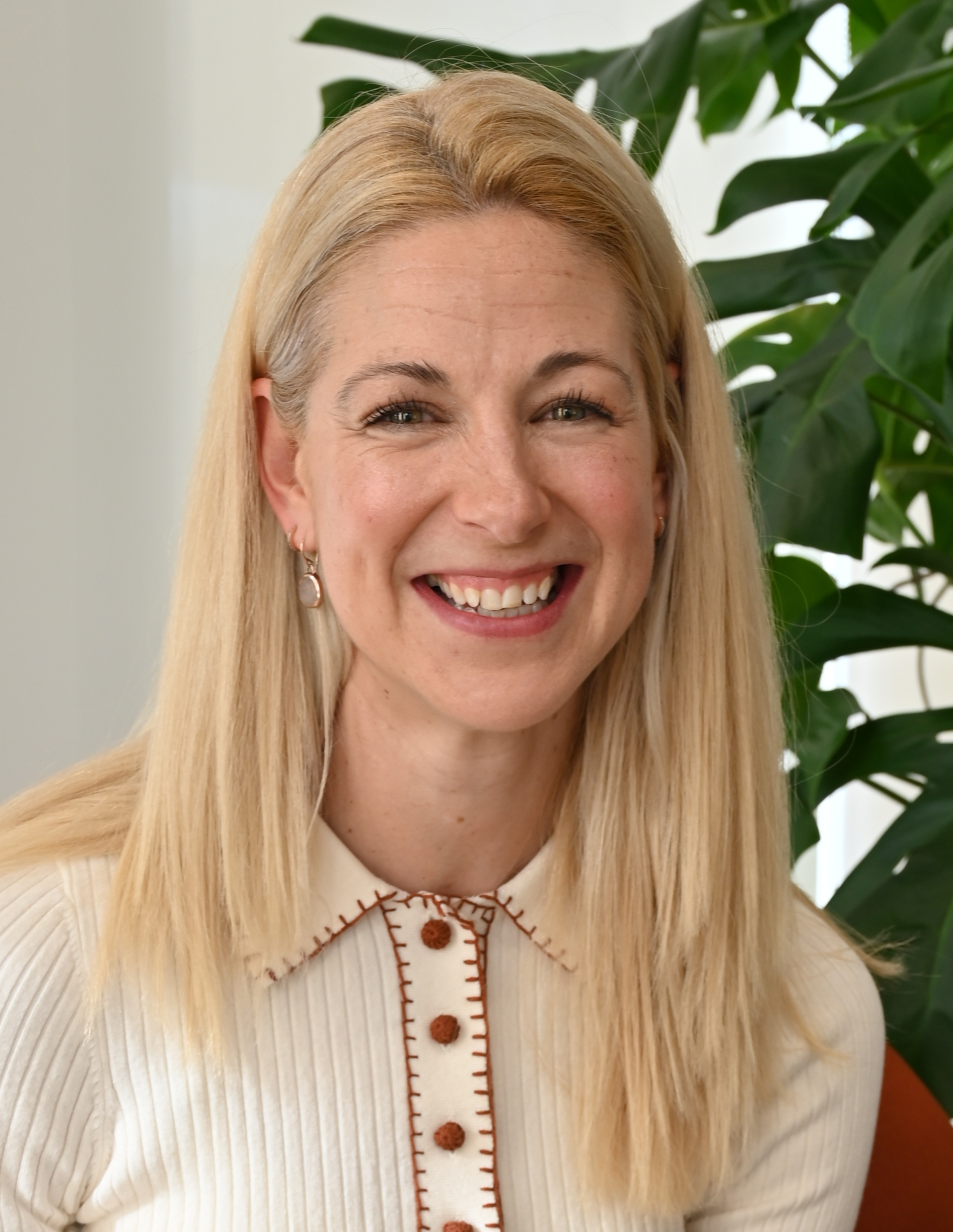 Smiling woman with long blonde hair wearing a green top standing indoors near a plant.