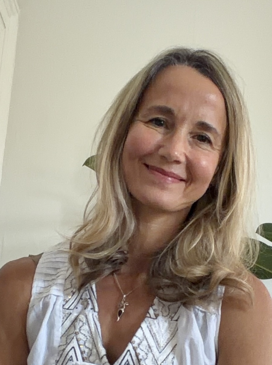 Smiling woman with long wavy blonde hair sitting indoors near a plant and beige wall panels.